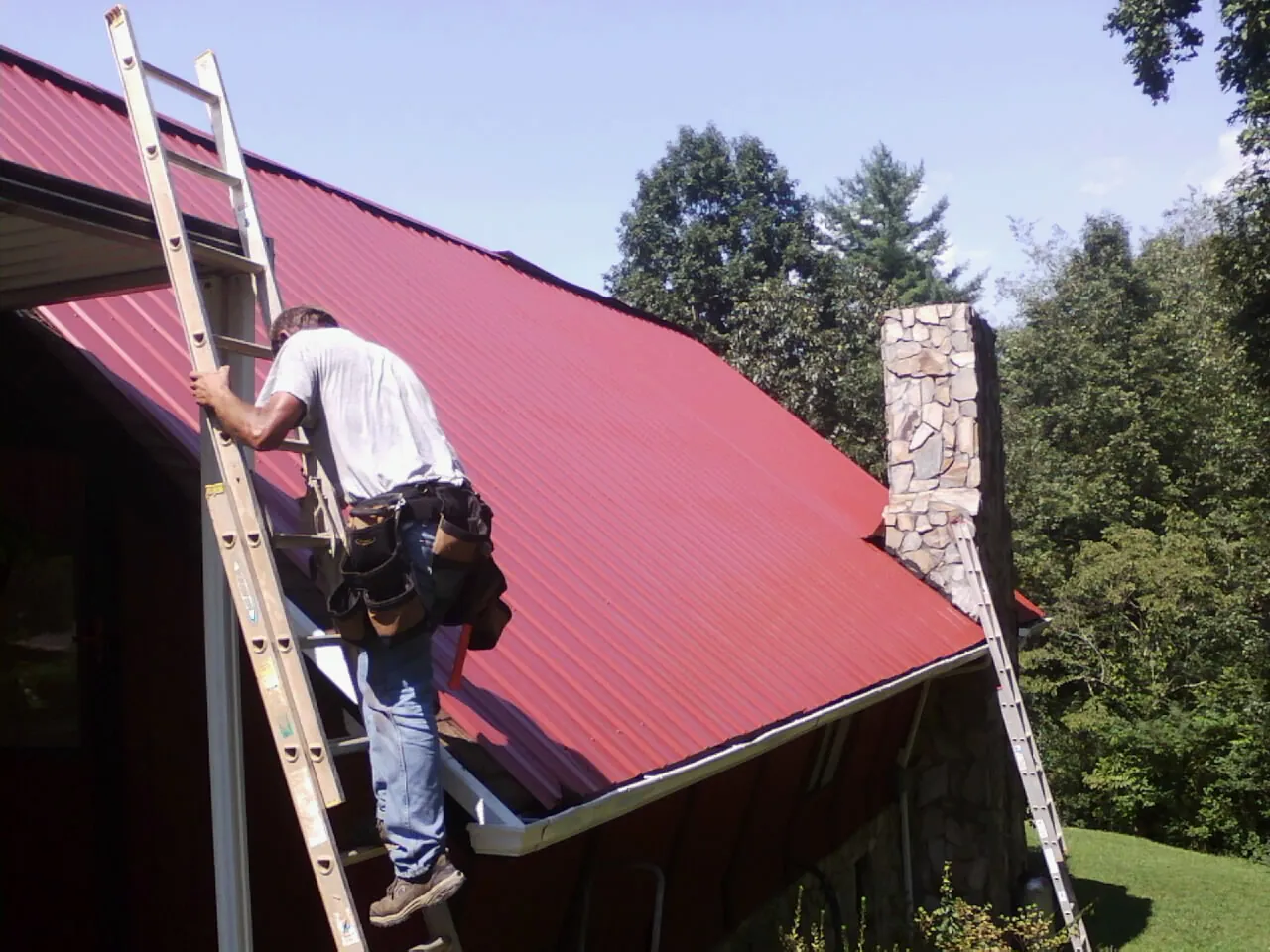 Roofer on a ladder beside a red metal roof and stone chimney — Soffit & Fascia Repair by Roofing Brothers in Port Jervis